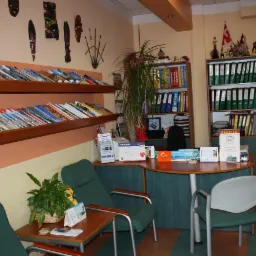 Wnętrze biura z półkami pełnymi broszur turystycznych, egzotyczne maski na ścianie, zielone fotele i biurko z materiałami reklamowymi.