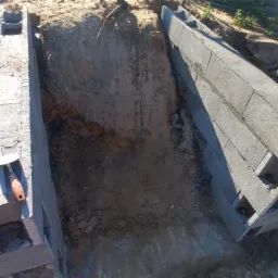 Wykop fundamentów z częściowo wzniesioną ścianą z szarych bloczków betonowych z pustakami, widoczne narzędzia murarskie i wykopana ziemia.