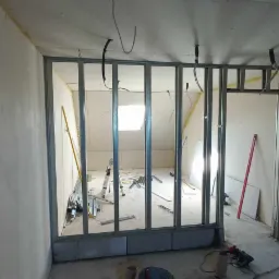 Wnętrze w trakcie tworzenia ścianki działowej z metalowych profili, widoczne narzędzia, resztki materiałów i instalacja elektryczna pod sufitem.