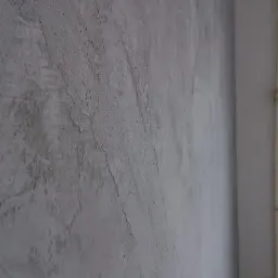 Szara, surowa ściana w trakcie nakładania gładzi, widoczne nierówności i faktura, sufit i róg ściany w tle.