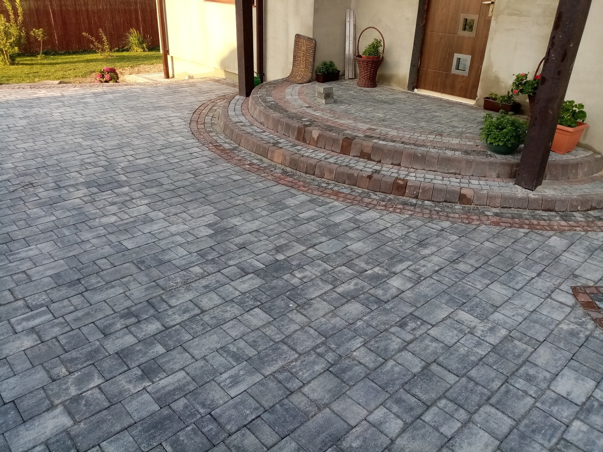 Szara kostka brukowa ułożona na podjeździe z obrzeżami z czerwonej kostki, prowadząca do wejścia z trzema stopniami, na których stoją doniczki z kwiatami.