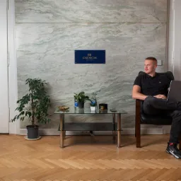 Mężczyzna siedzi w fotelu z laptopem na tle marmurowej ściany z logo firmy Esencja Nieruchomości, obok stolik kawowy z roślinami i butelkami wody.