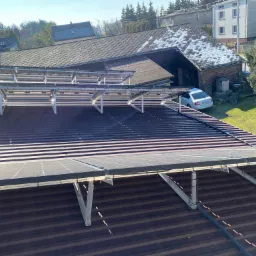 Panele słoneczne zamontowane na brązowym dachu blaszanym, widok z góry na domy i ogród w tle.