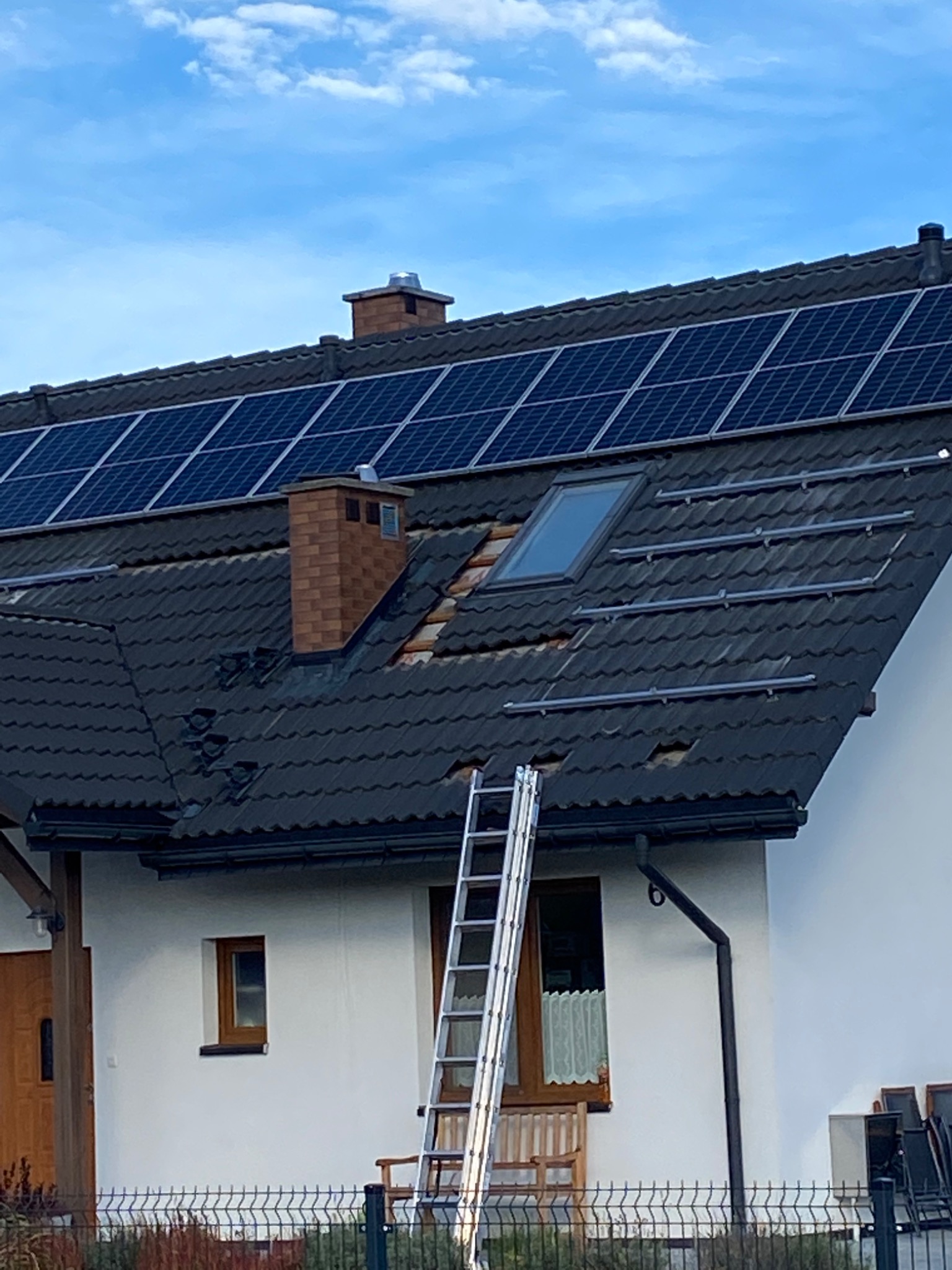 Montaż paneli słonecznych na dachu domu jednorodzinnego, widoczne częściowo zamontowane panele, aluminiowe szyny montażowe, drabina oparta o ścianę budynku, dachówka w kolorze grafitowym, ceglany...