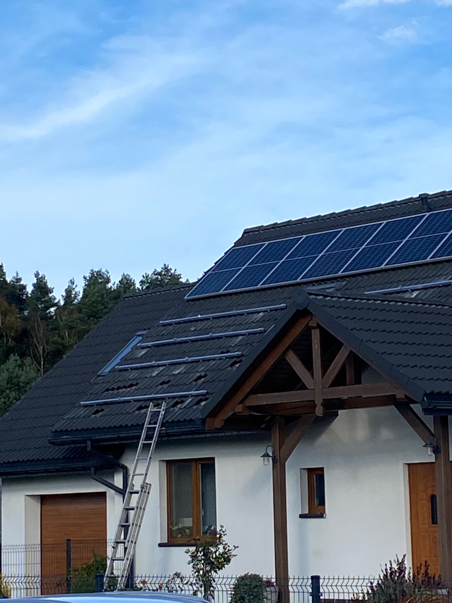 Montaż paneli słonecznych na dachu domu jednorodzinnego z czarną dachówką, widoczne szyny montażowe i drabina oparta o budynek. Część paneli już zamontowana.