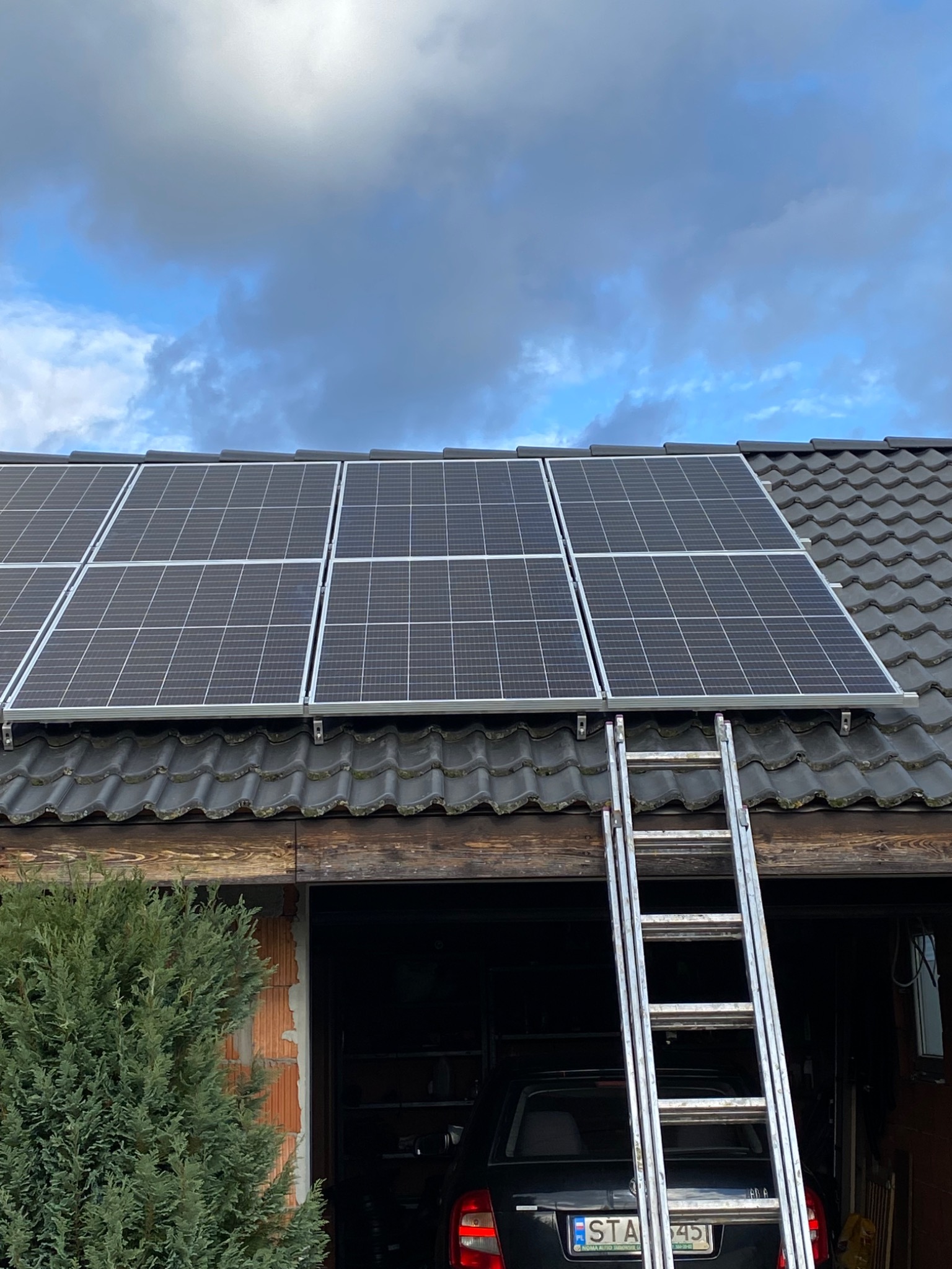 Panele fotowoltaiczne na dachu domu w Tąpkowicach, z rozstawioną aluminiową drabiną opartą o dach, widok z dołu na tle pochmurnego nieba.