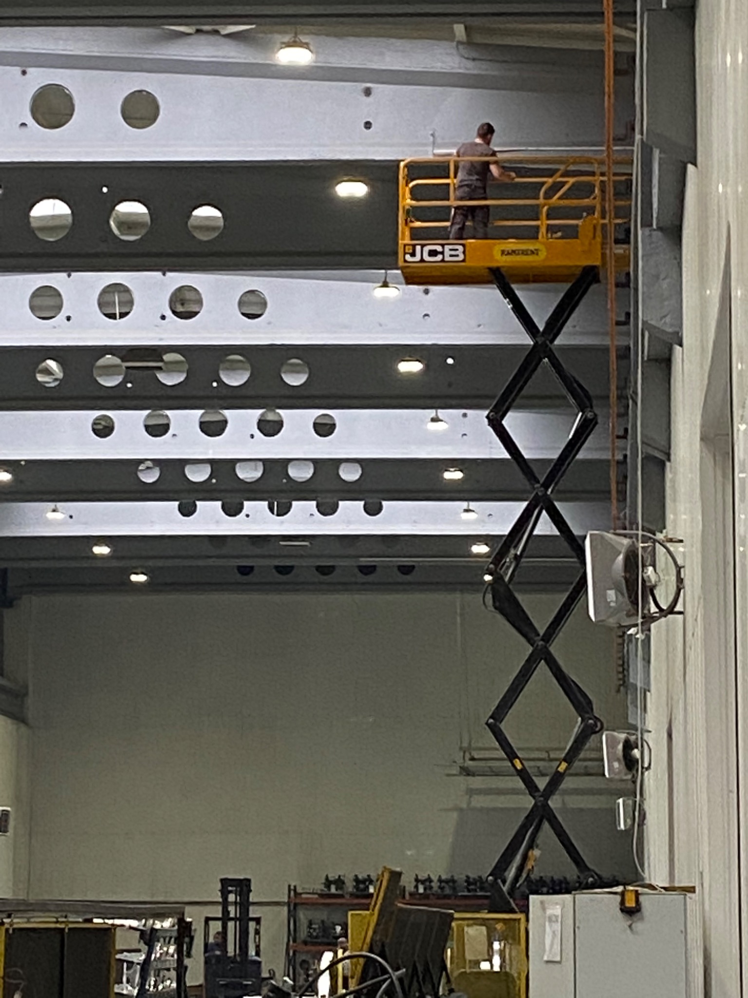 Pracownik na podnośniku nożycowym JCB, wykonujący prace na wysokości przy konstrukcji sufitu z widocznymi otworami, w hali przemysłowej.
