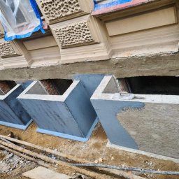 Firma budowlana NIK MAR Artur Jung - Wykonane betonowe obudowy okien piwnicznych, częściowo pomalowane na szaro, z widoczną strukturą szalunku na jednej ze ścianek; w tle fragment elewacji budynku z dekoracyjnymi elementami.