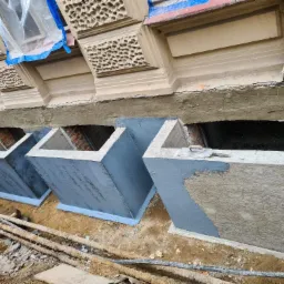 Wykonane betonowe obudowy okien piwnicznych, częściowo pomalowane na szaro, z widoczną strukturą szalunku na jednej ze ścianek; w tle fragment elewacji budynku z dekoracyjnymi elementami.