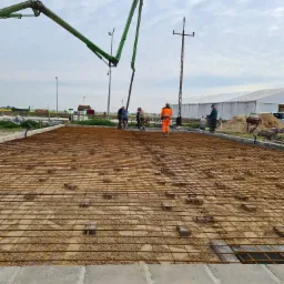 Przygotowanie zbrojenia pod wylewkę fundamentową z użyciem pompy do betonu, widoczni pracownicy w odzieży roboczej, teren budowy.