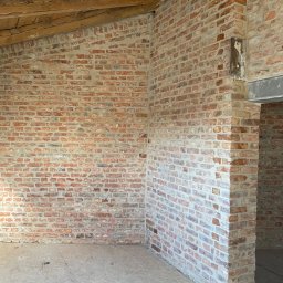 Firma budowlana NIK MAR Artur Jung - Wnętrze w trakcie budowy z czerwoną cegłą, drewnianym stropem i surową podłogą z płyty OSB.