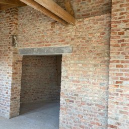 Firma budowlana NIK MAR Artur Jung - Wnętrze domu w trakcie budowy z widocznymi ścianami z czerwonej cegły, drewnianymi belkami stropowymi i surową podłogą, ukazujące etap murowania ścian i przygotowania do dalszych prac wykończeniowych.