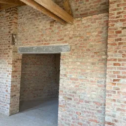 Wnętrze domu w trakcie budowy z widocznymi ścianami z czerwonej cegły, drewnianymi belkami stropowymi i surową podłogą, ukazujące etap murowania ścian i przygotowania do dalszych prac wykończeniowych.