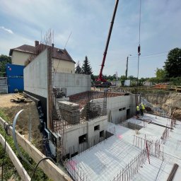 Firma budowlana NIK MAR Artur Jung - Wykonywanie fundamentów budynku żelbetowego z widocznym zbrojeniem, deskowaniem i pracującym żurawiem na placu budowy w słoneczny dzień.