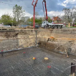 Zbrojenie fundamentu z widoczną pompą do betonu Putzmeister na placu budowy, w tle budynki mieszkalne i pochmurne niebo.