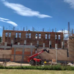 Wznoszony budynek z czerwonej cegły i pustaków, z widocznym rusztowaniem, elementami zbrojenia i podnośnikiem teleskopowym Manitou na pierwszym planie.