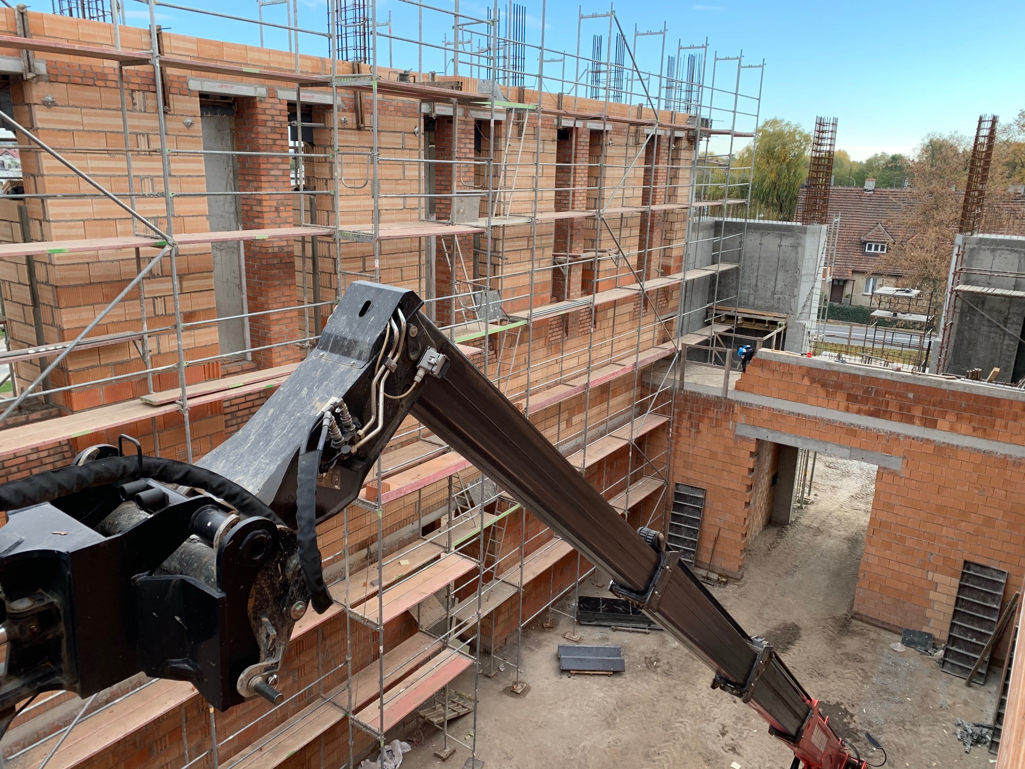 Wznoszenie dwukondygnacyjnego budynku z czerwonej cegły ceramicznej, widok z góry na rusztowania i dźwig z chwytakiem do przenoszenia bloczków, w tle zieleń i zabudowa jednorodzinna.