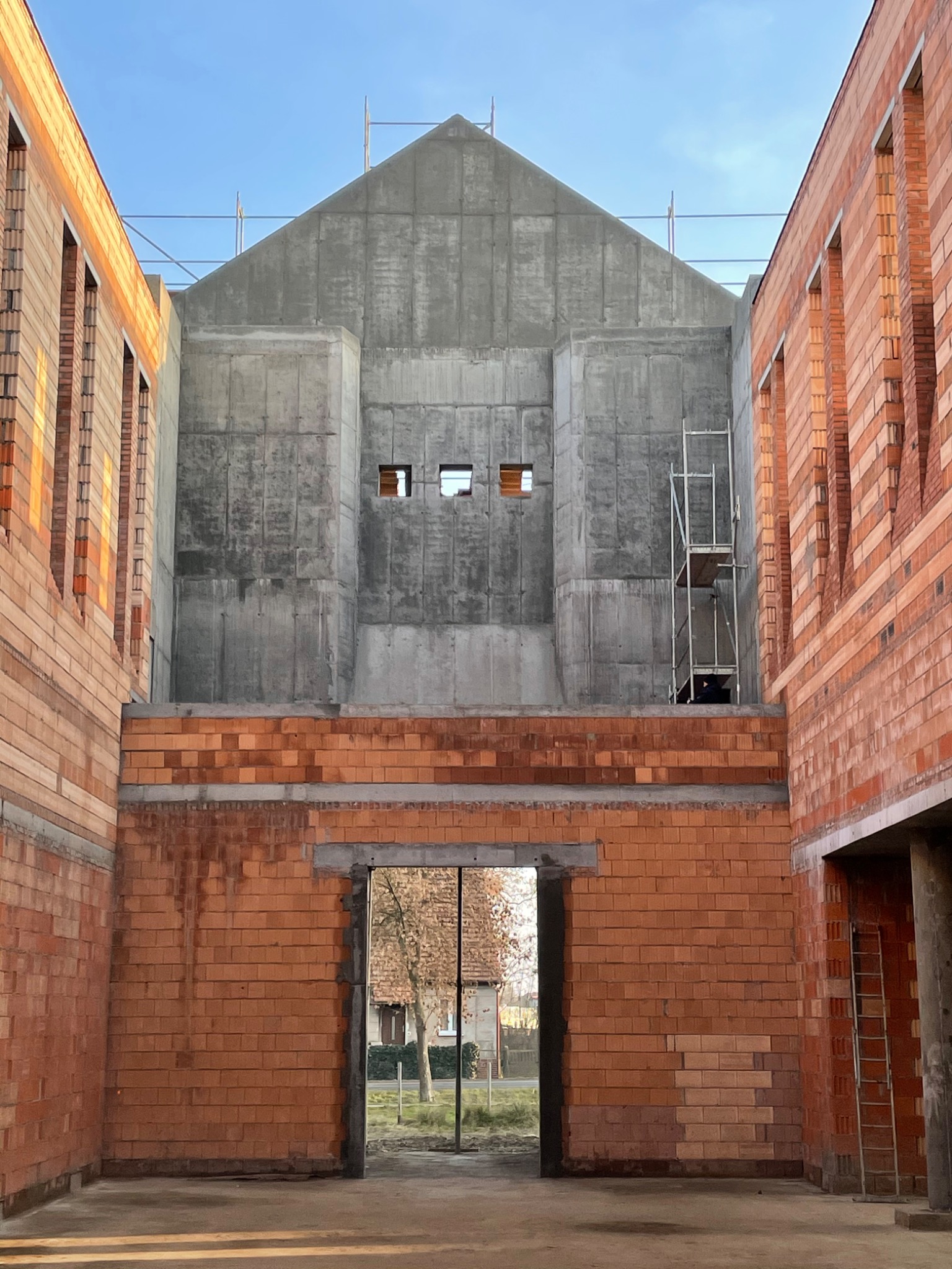 Surowy stan budynku: ściany z czerwonej cegły klinkierowej w trakcie wznoszenia, centralnie betonowa konstrukcja z małymi oknami, widoczne rusztowanie i drabina, w tle drzewa.