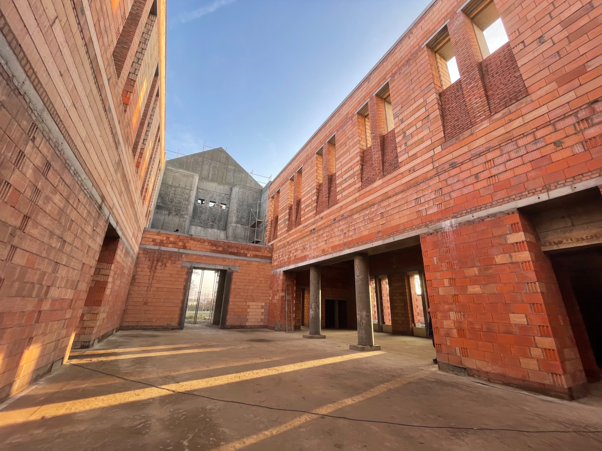Wnętrze budynku w trakcie budowy z czerwonej cegły, widoczne betonowe kolumny i otwory okienne bez ram, w tle budynek z betonu, wczesne stadium budowy.