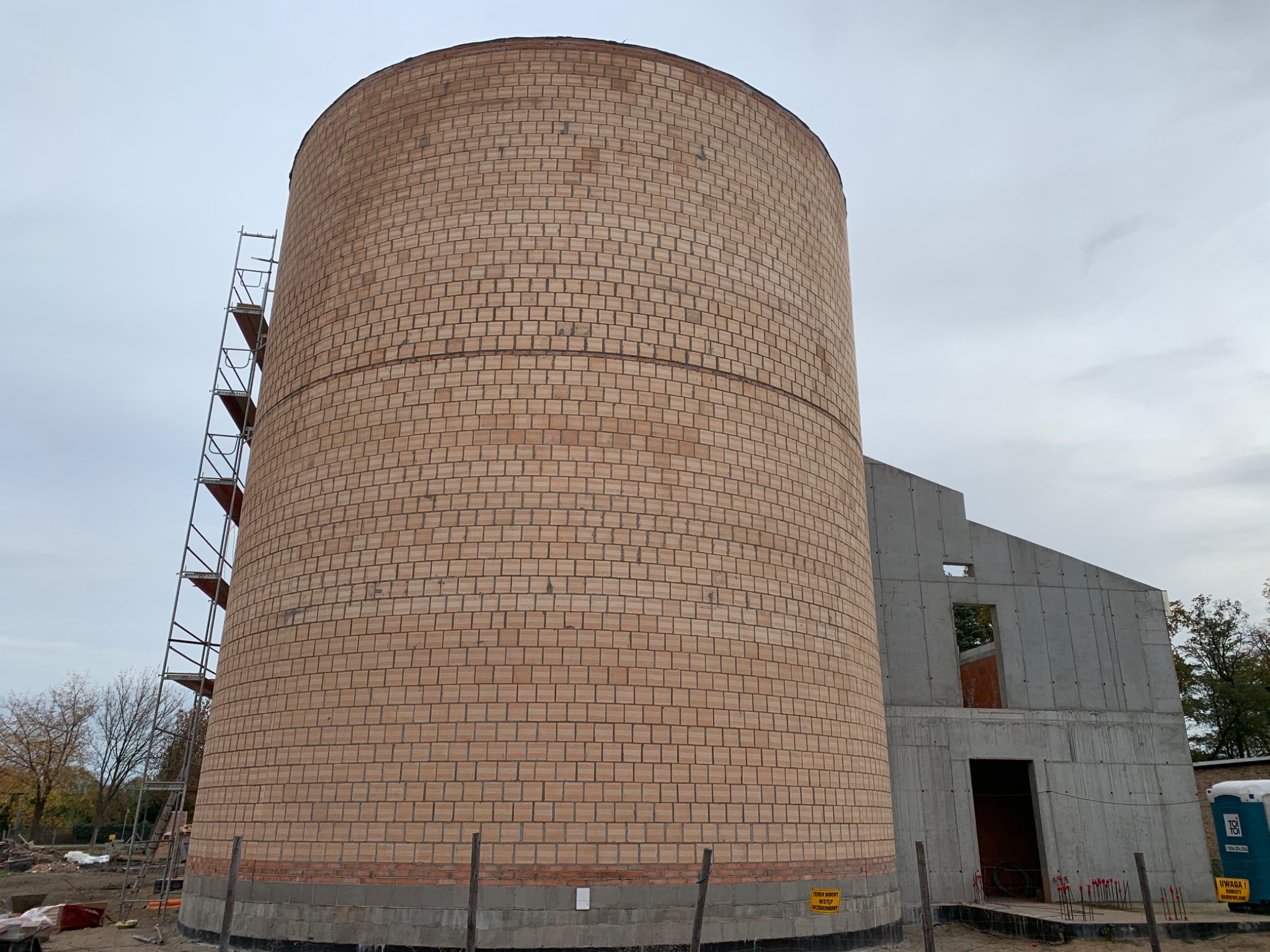 Cylindryczna konstrukcja z cegły na etapie budowy, z widocznymi rusztowaniami i fragmentem budynku żelbetowego w tle, teren budowy z oznaczeniami ostrzegawczymi.