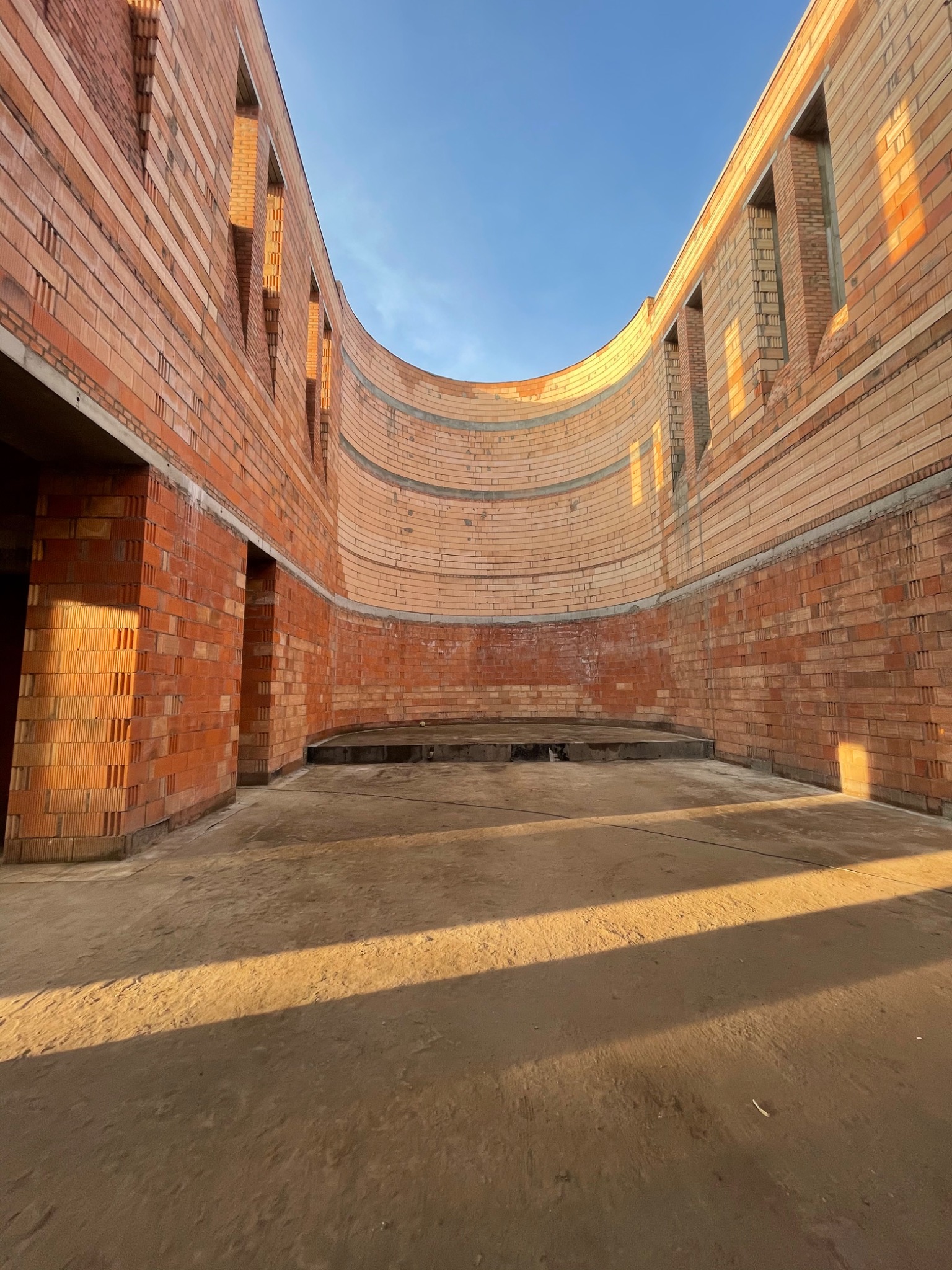 Wnętrze budynku w trakcie budowy z czerwonej cegły, widoczne ściany z otworami okiennymi i drzwiowymi, surowy betonowy podkład, wpadające promienie słoneczne tworzą długie cienie.