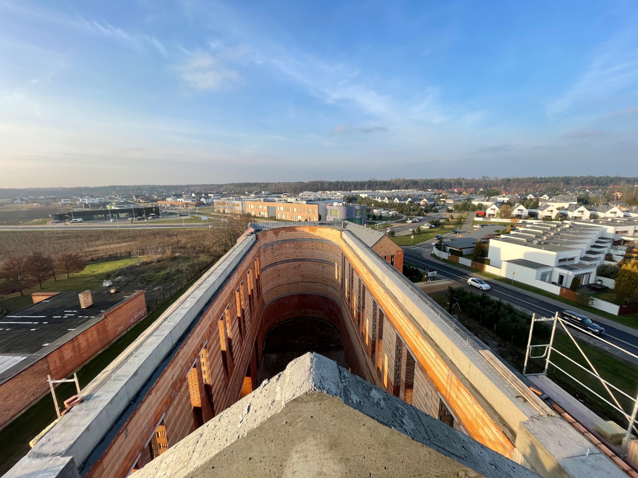 Wysoki kąt widzenia na budynek w trakcie budowy z czerwonej cegły z widocznymi elementami betonowymi i rusztowaniem, na tle miejskiego krajobrazu z niebem pokrytym chmurami.