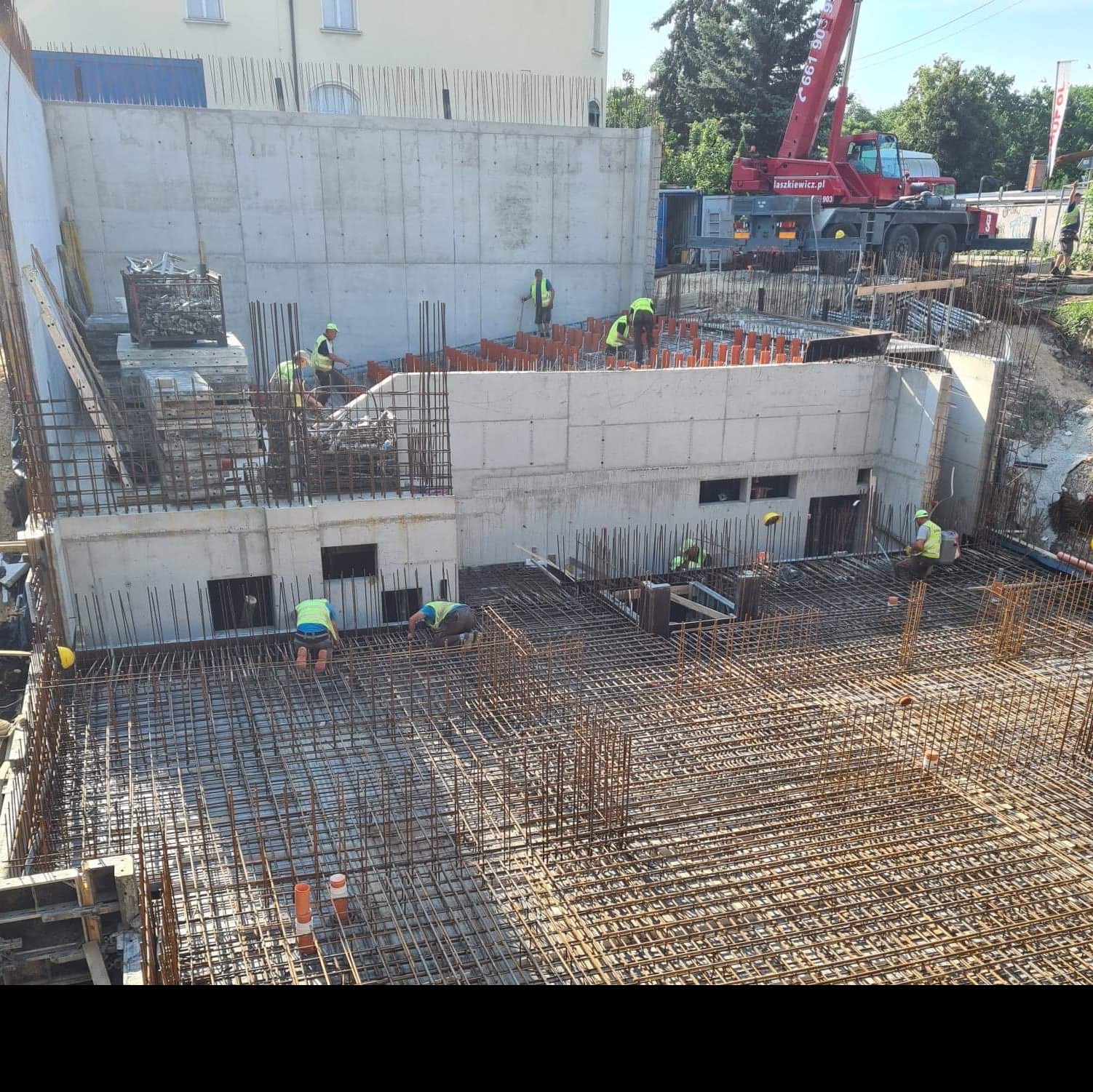 Wielopoziomowy plac budowy z widoczną konstrukcją żelbetową w trakcie wznoszenia, zbrojenia stalowe na różnych poziomach, pracownicy w kamizelkach odblaskowych i żuraw wieżowy w tle.