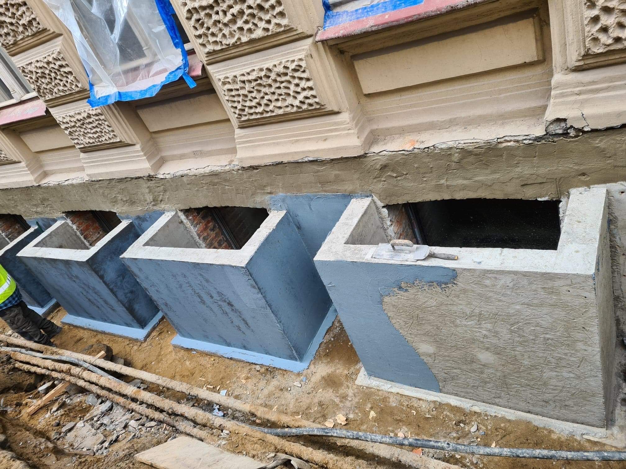 Wykonane betonowe obudowy okien piwnicznych, częściowo pomalowane na szaro, z widoczną strukturą szalunku na jednej ze ścianek; w tle fragment elewacji budynku z dekoracyjnymi elementami.