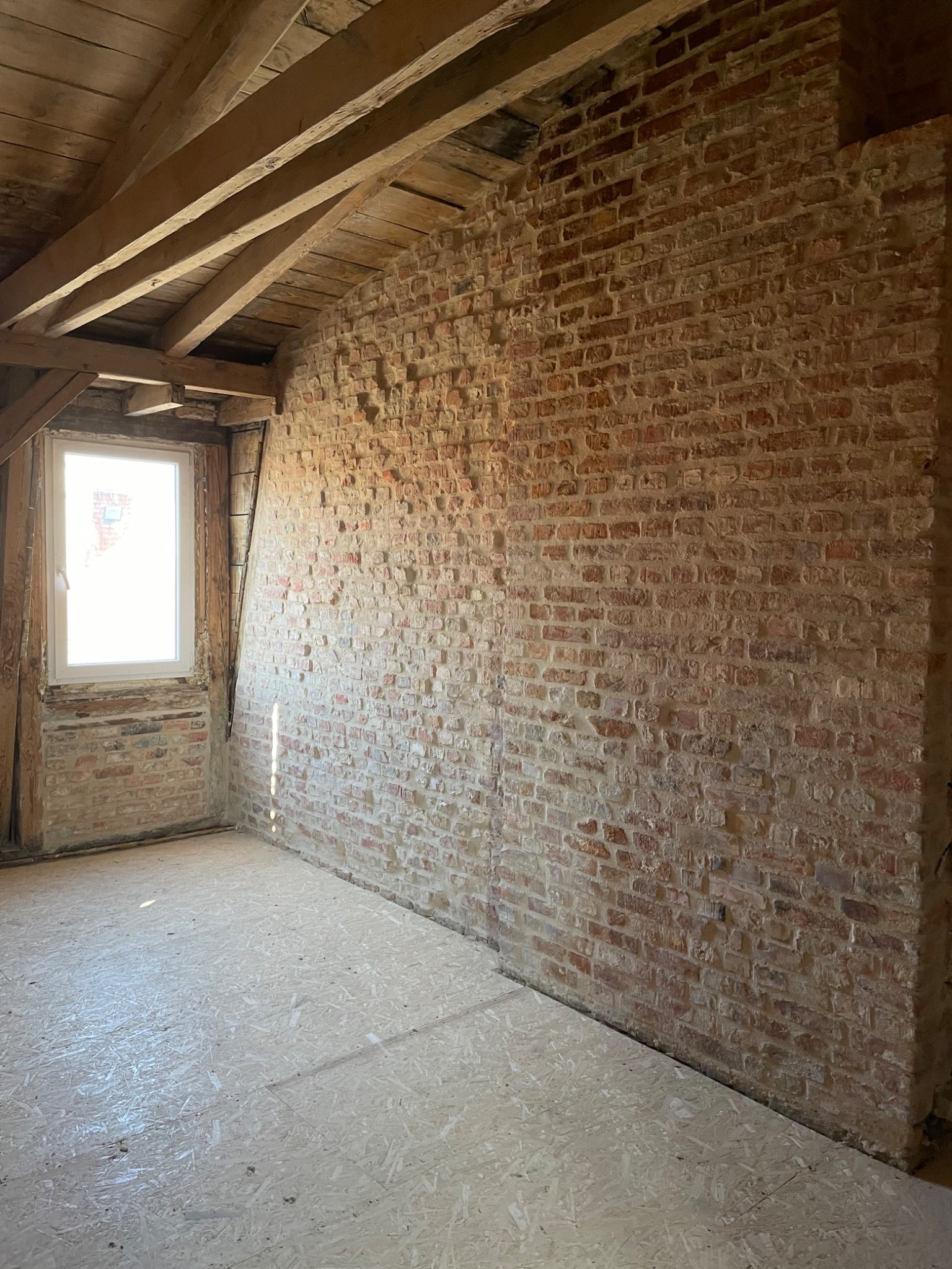Wnętrze poddasza w trakcie remontu: surowa ściana z czerwonej cegły z widocznymi śladami zaprawy, drewniana konstrukcja dachu, nowe okno z białymi ramami i podłoga wyłożona płytami OSB.