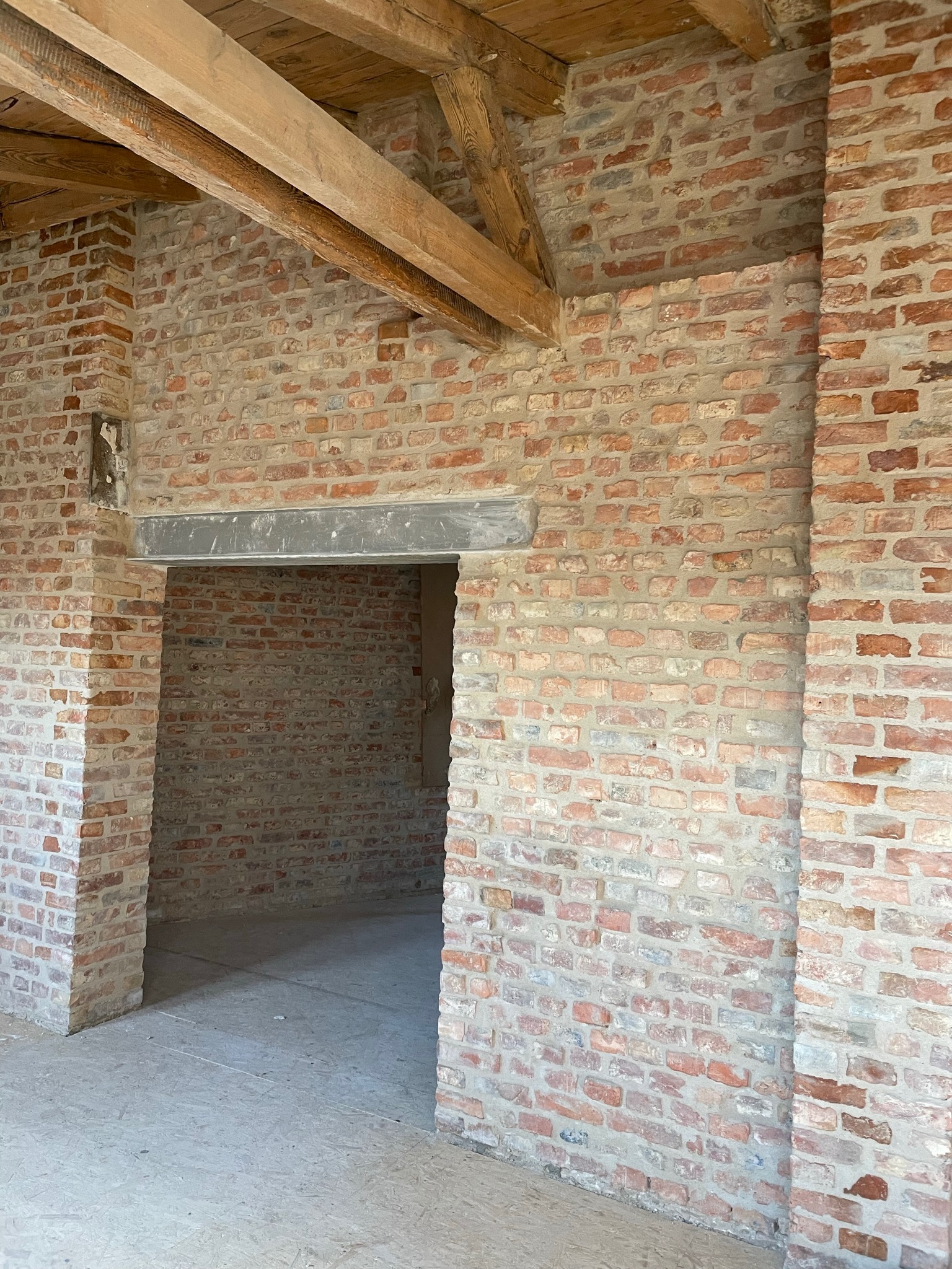 Wnętrze domu w trakcie budowy z widocznymi ścianami z czerwonej cegły, drewnianymi belkami stropowymi i surową podłogą, ukazujące etap murowania ścian i przygotowania do dalszych prac wykończeniowych.