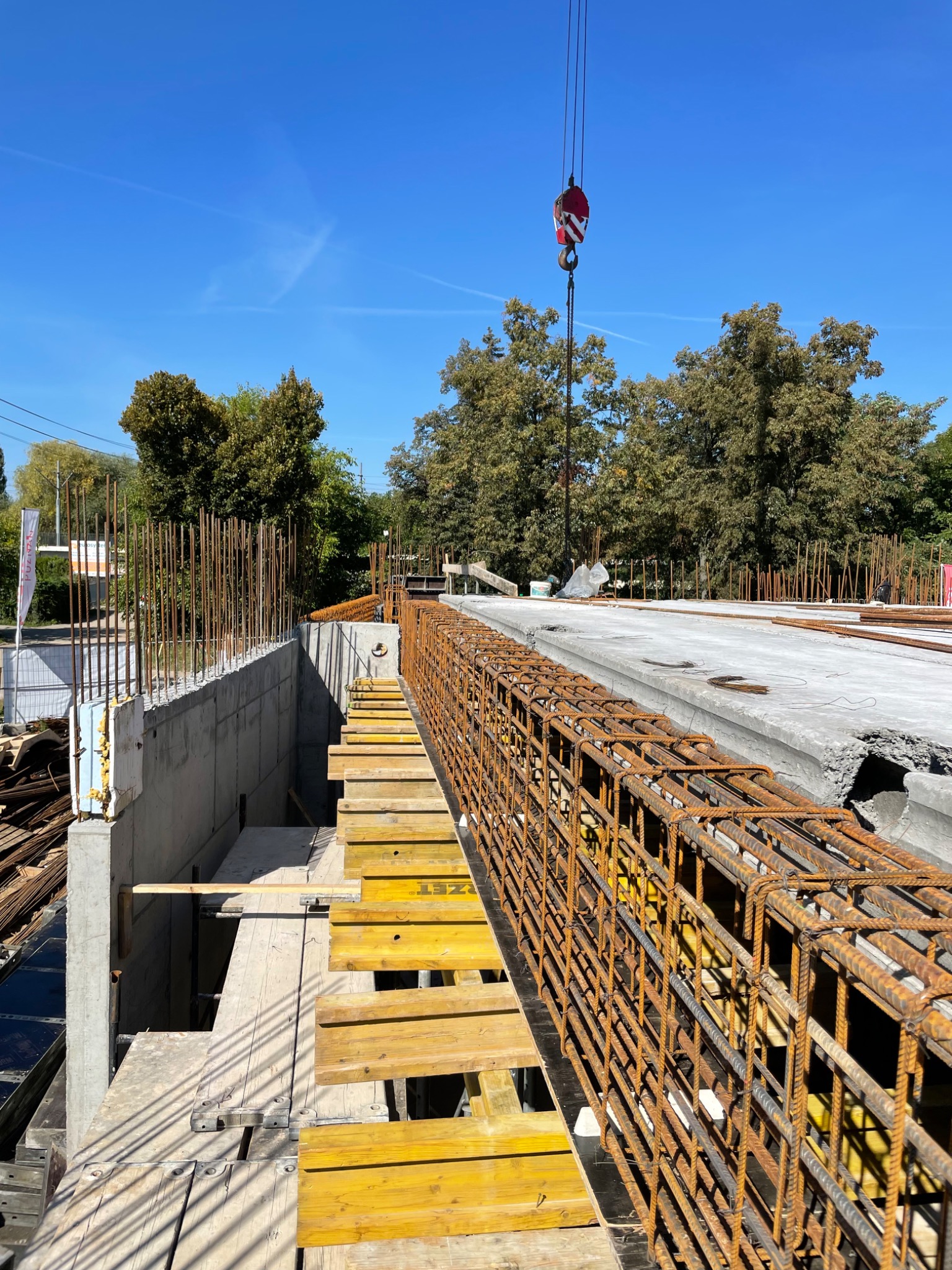 Żelbetowa konstrukcja w trakcie budowy, z widocznym zbrojeniem z prętów stalowych, szalunkiem z żółtych desek, betonowymi ścianami i dźwigiem budowlanym na tle błękitnego nieba i drzew.