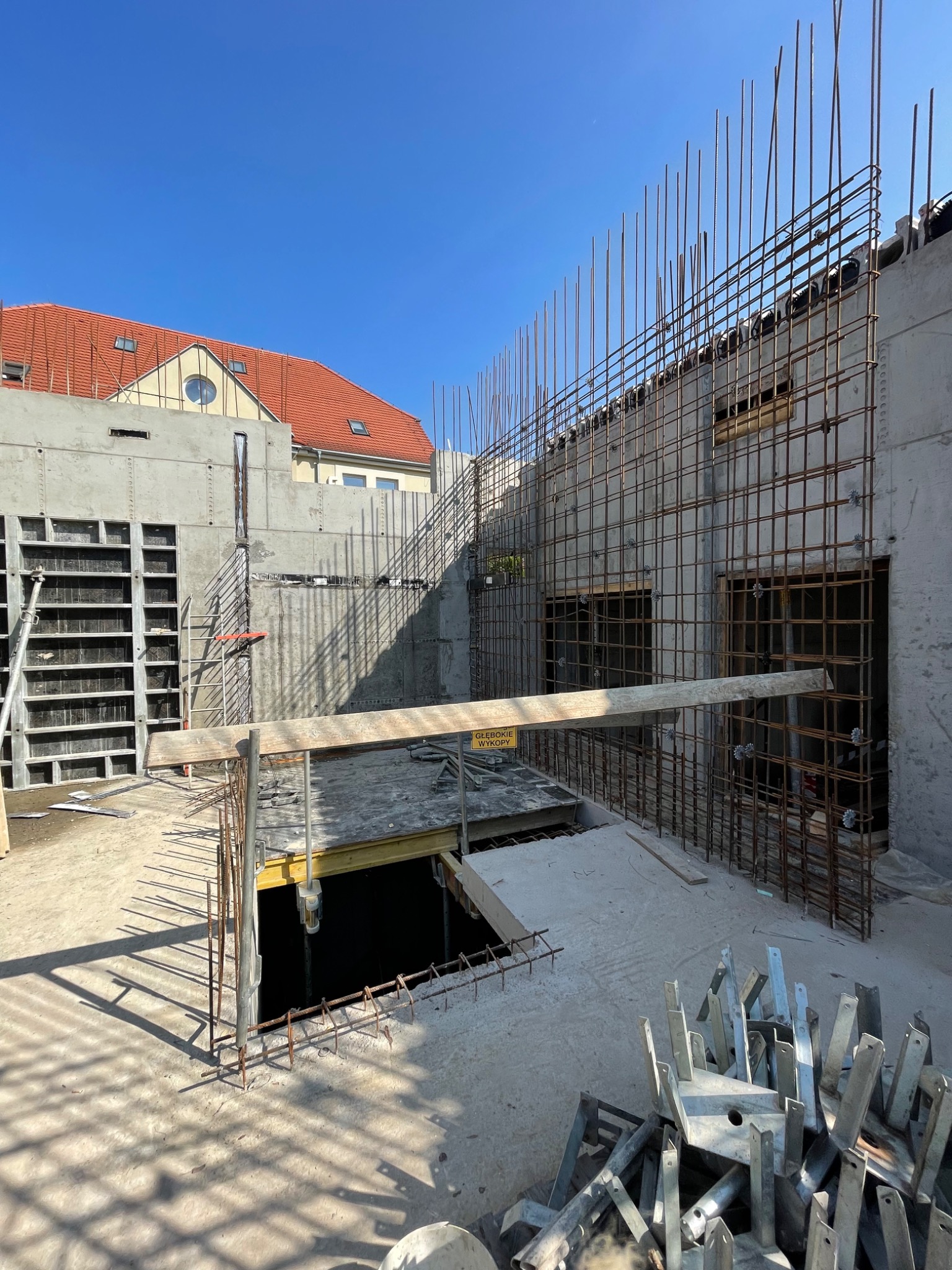 Wznoszenie budynku w technologii szkieletu żelbetowego, widoczne zbrojenia ścian z otworami okiennymi, szalunki na ścianie obok i elementy konstrukcyjne na placu budowy w słoneczny dzień.