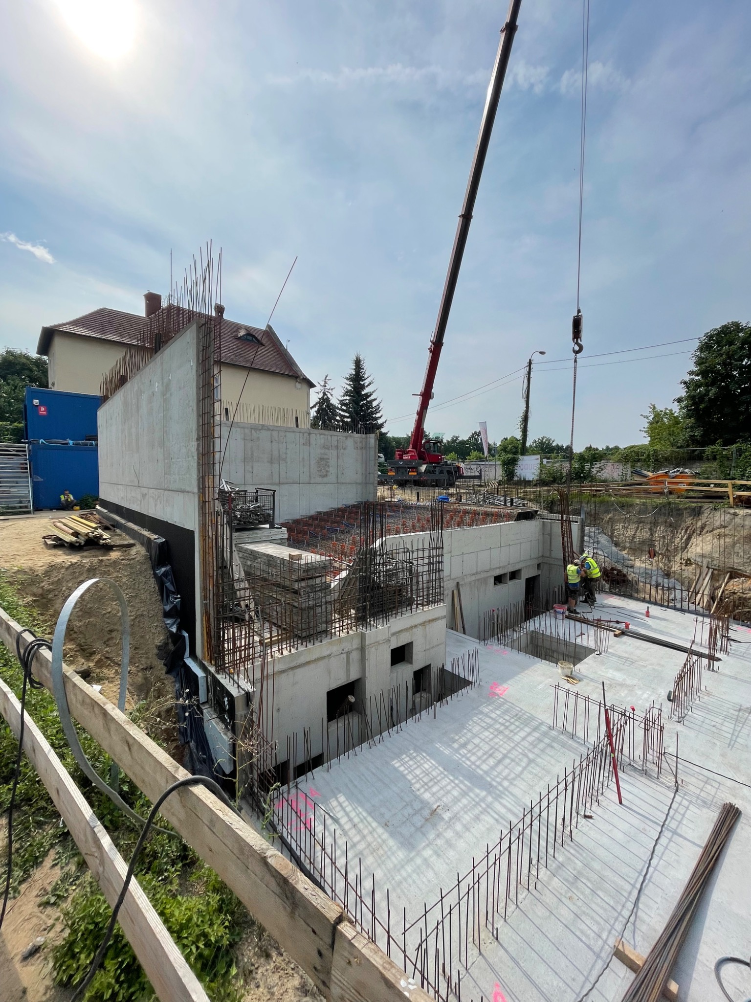 Wykonywanie fundamentów budynku żelbetowego z widocznym zbrojeniem, deskowaniem i pracującym żurawiem na placu budowy w słoneczny dzień.