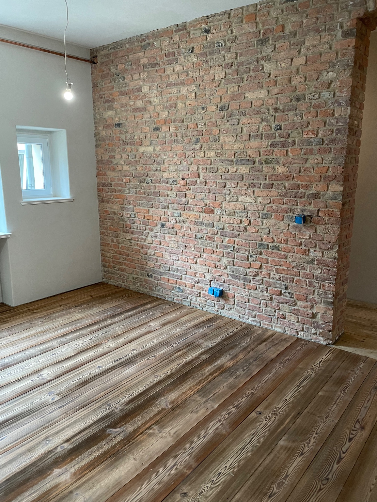 Wnętrze z odsłoniętą ścianą z czerwonej cegły, drewnianą podłogą z widocznym usłojeniem i niebieskimi puszkami elektrycznymi na ścianie.