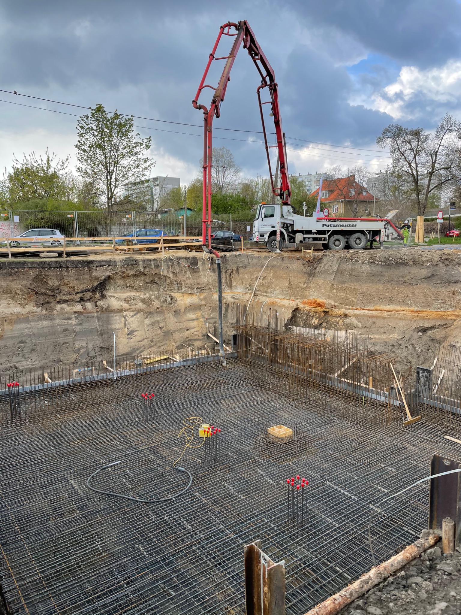 Zbrojenie fundamentu z widoczną pompą do betonu Putzmeister na placu budowy, w tle budynki mieszkalne i pochmurne niebo.