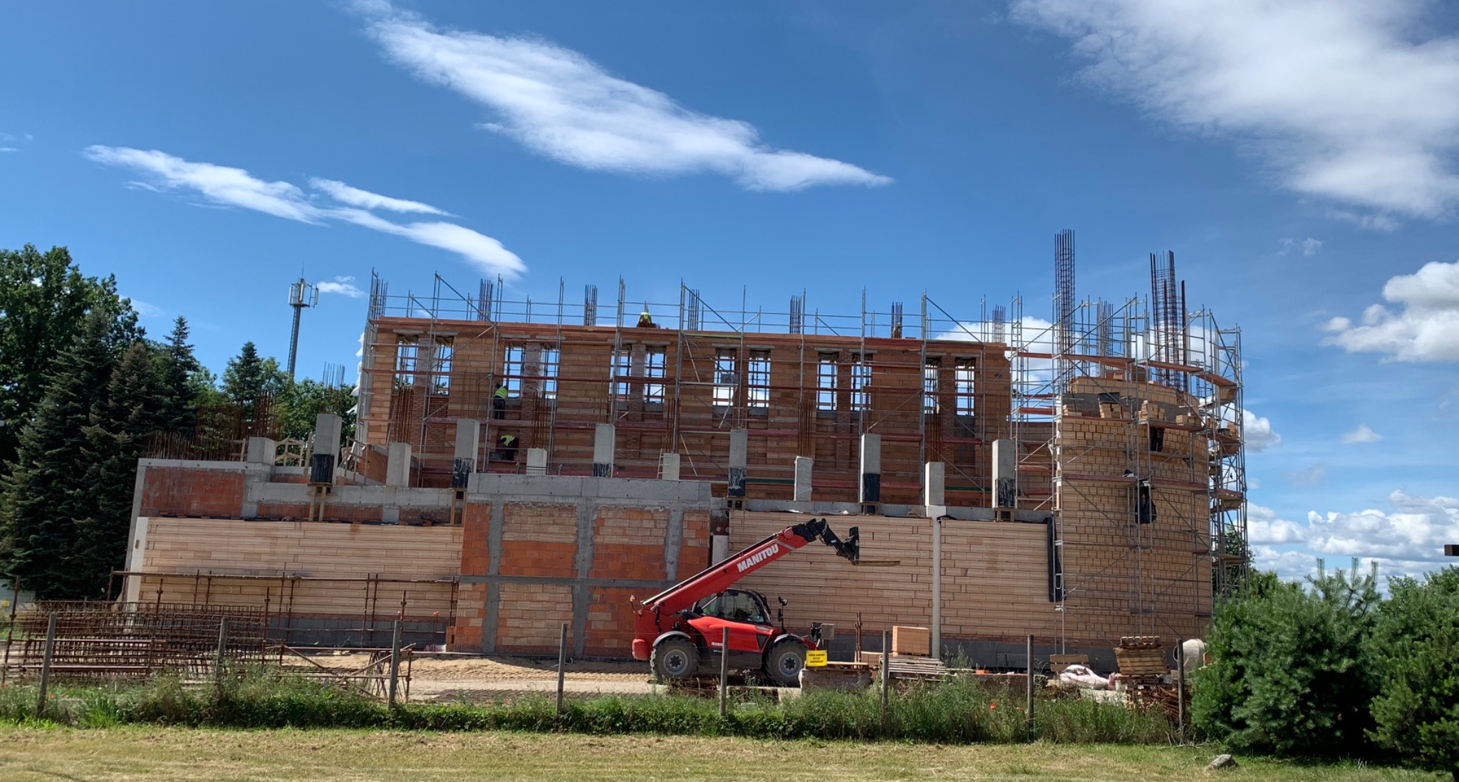 Wznoszony budynek z czerwonej cegły i pustaków, z widocznym rusztowaniem, elementami zbrojenia i podnośnikiem teleskopowym Manitou na pierwszym planie.