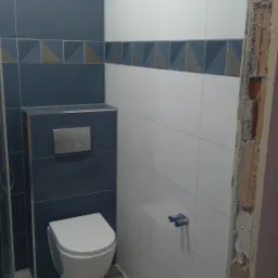 Świeżo wyremontowana łazienka z wiszącą toaletą, szarymi i białymi płytkami oraz dekoracyjnym pasem z geometrycznym wzorem, widoczny fragment surowego ościeża drzwi.