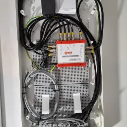 Wewnętrzna skrzynka rozdzielcza z zamontowanym multiswitchem Corab, widoczne kable koncentryczne, ethernetowe i zasilające, starannie ułożone wewnątrz obudowy.