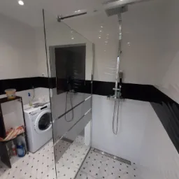 Nowoczesna kabina prysznicowa typu walk-in z szklaną ścianką, deszczownicą i odpływem liniowym w wykończonej łazience z biało-czarnymi płytkami i pralką w tle.