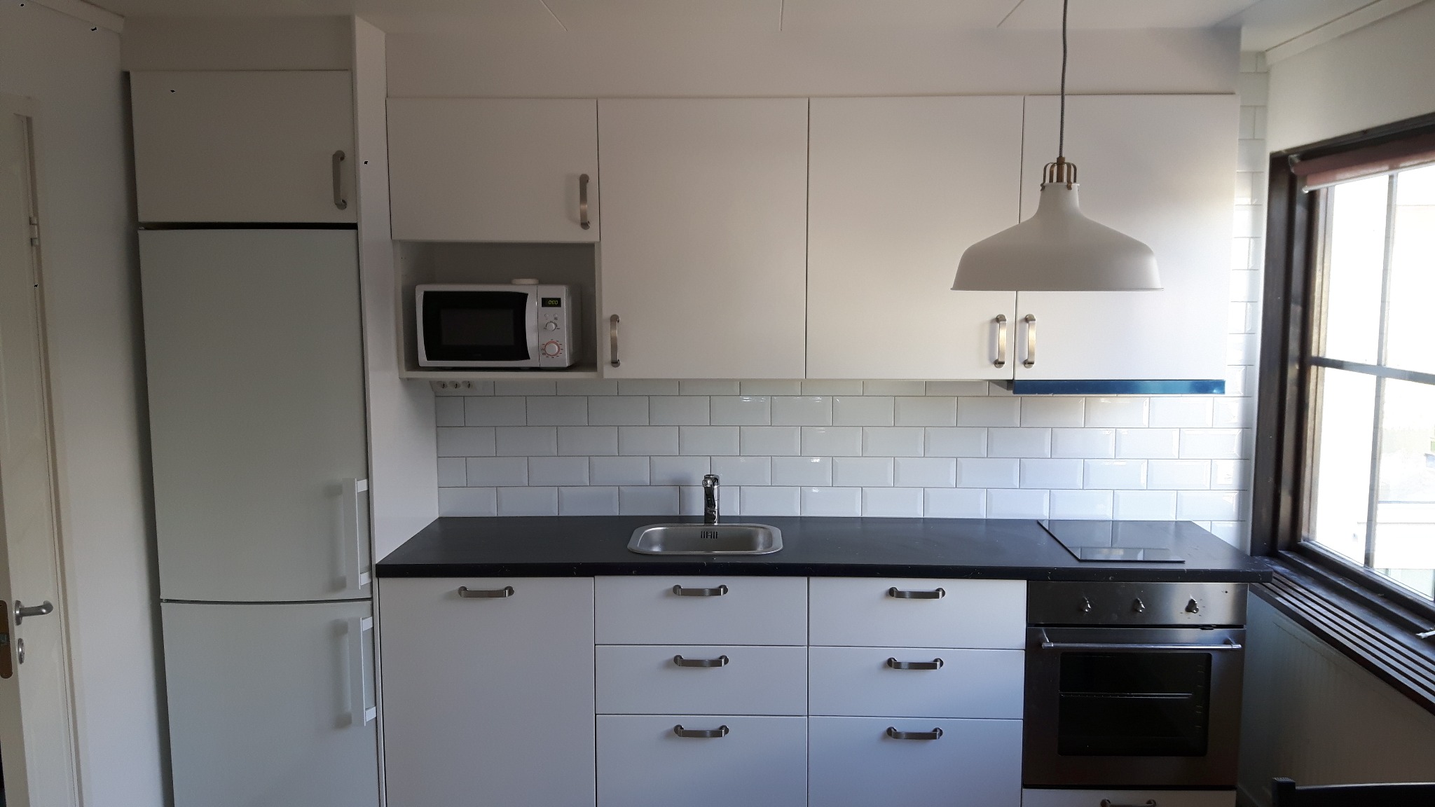 Nowoczesna, jasna kuchnia z białymi szafkami, czarnym blatem, białymi płytkami na ścianie i oświetleniem punktowym, widok na kuchenkę i zlew