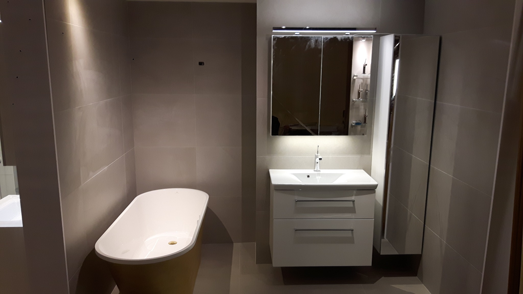 Ukończona łazienka z beżowymi płytkami, wolnostojąca wanna z żółtą obudową, biała szafka z umywalką i lustrem z oświetleniem LED, oraz szafka wisząca.