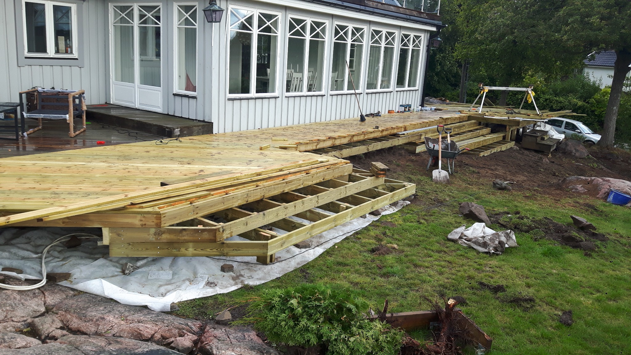 Budowa drewnianego tarasu przy domu z białymi oknami, widoczne schody w trakcie montażu, narzędzia pracy i przygotowane deski.