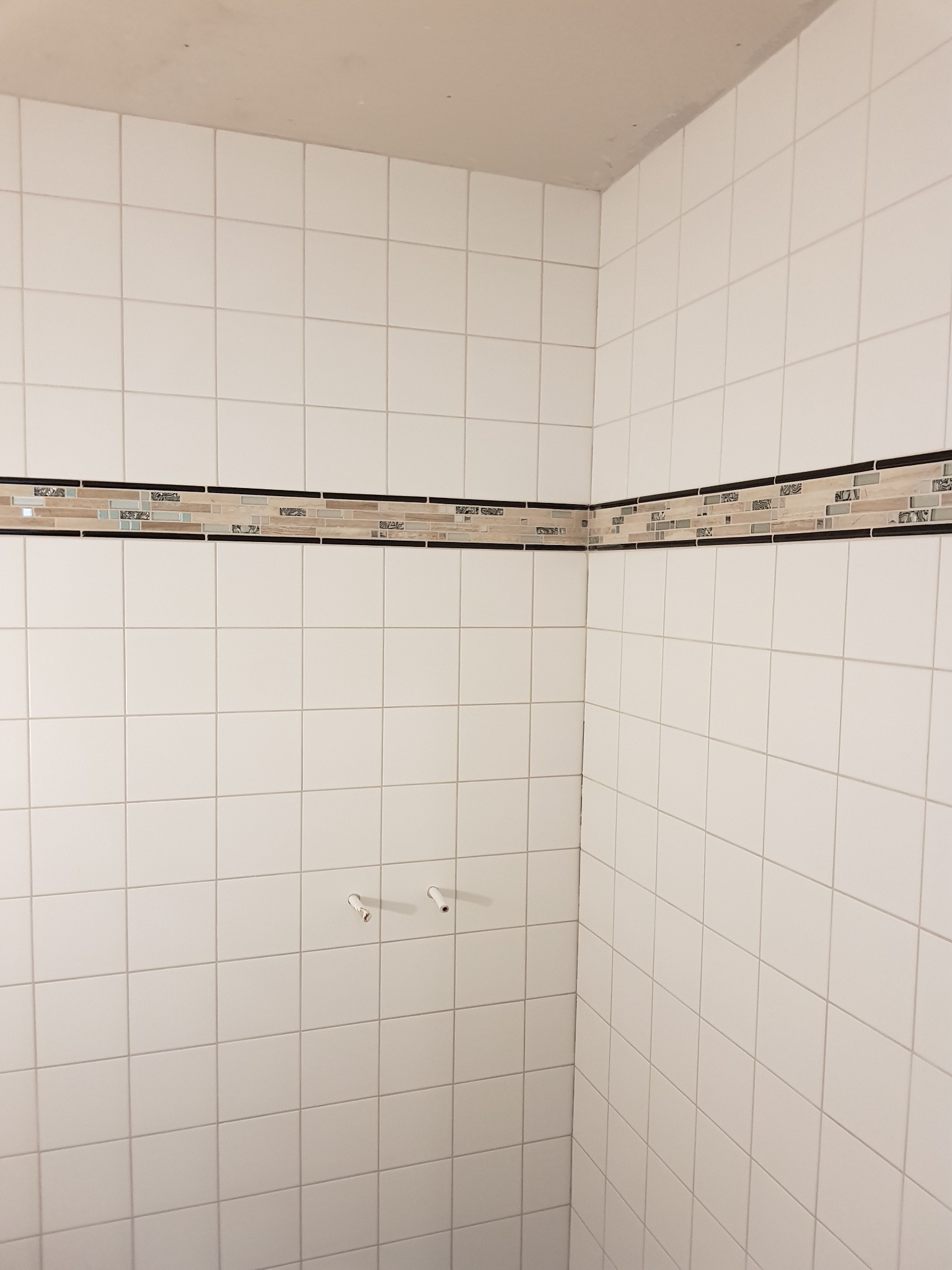 Wnętrze łazienki w trakcie prac: białe, kwadratowe płytki ceramiczne na ścianach z dekoracyjnym pasem mozaiki oraz wystającymi rurami instalacji wodnej.