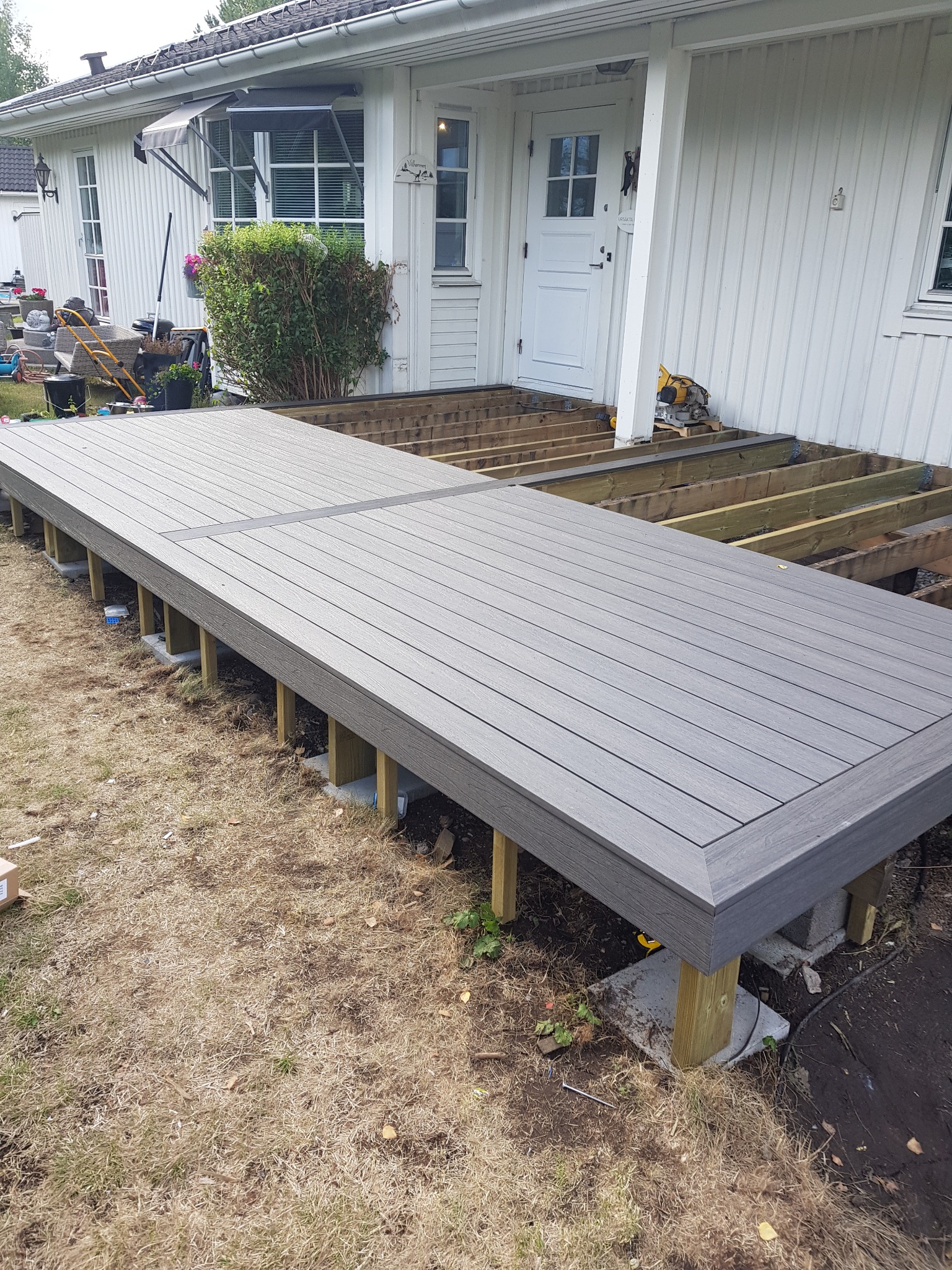Budowa szarego tarasu kompozytowego przy białym domu z widoczną konstrukcją nośną i narzędziami, trawa wokół.