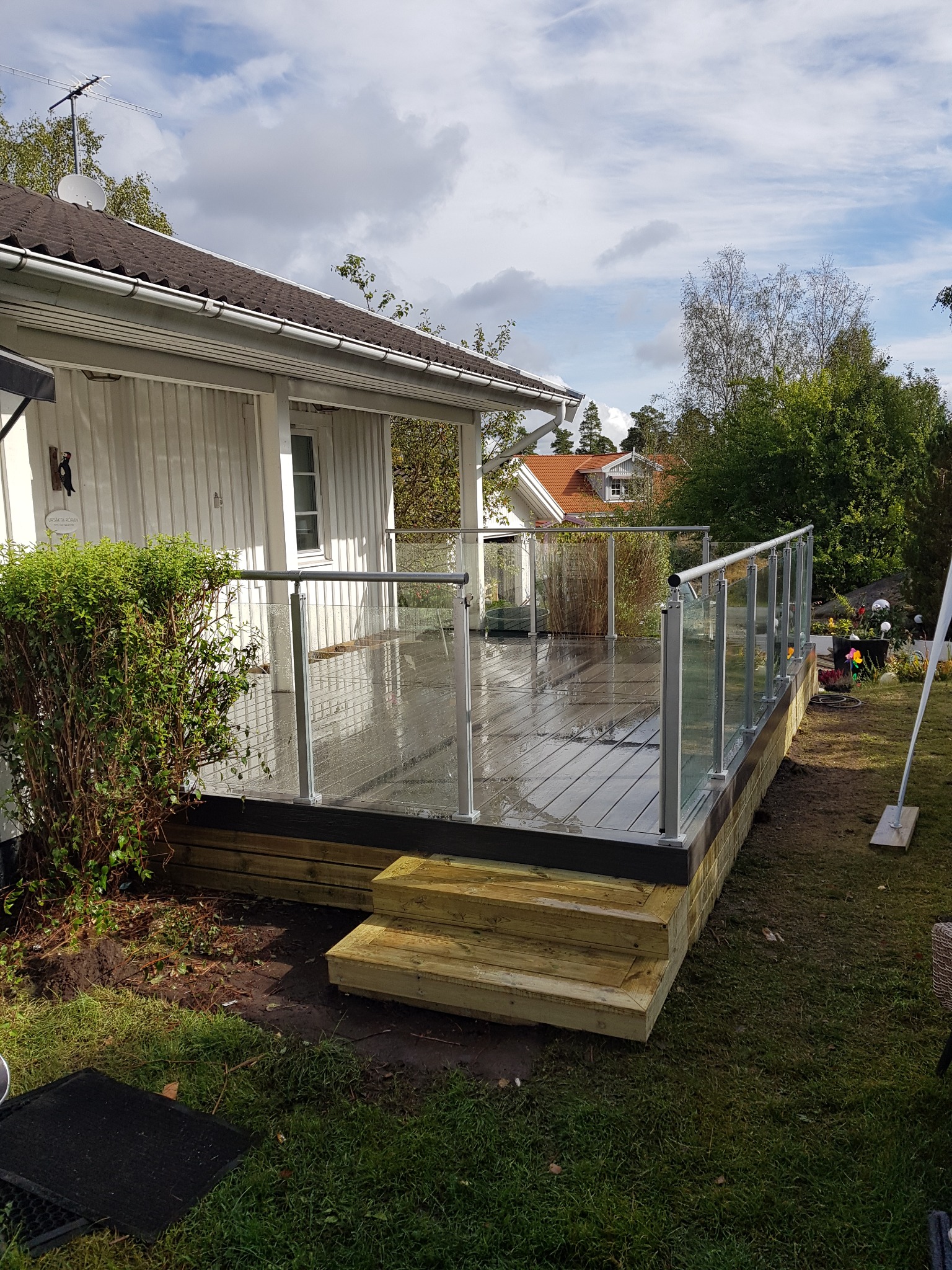 Nowoczesny taras z drewnianą podłogą i szklanymi balustradami, widok z zewnątrz na tle białego domu z brązowym dachem i zieleni.