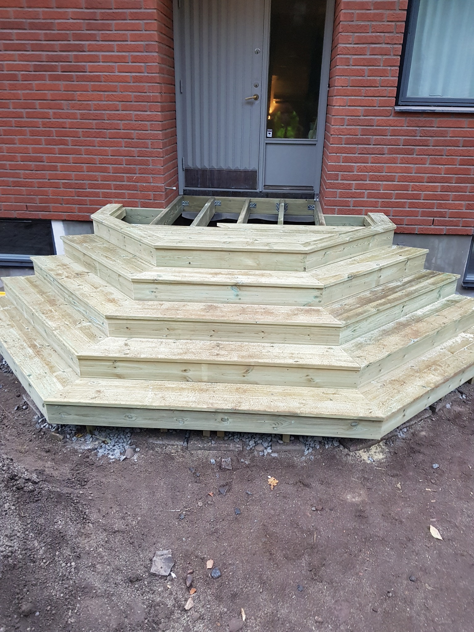 Nowe, drewniane schody wejściowe z jasnego drewna, prowadzące do drzwi wejściowych z ceglaną ścianą w tle, widoczne elementy konstrukcyjne pod schodami.