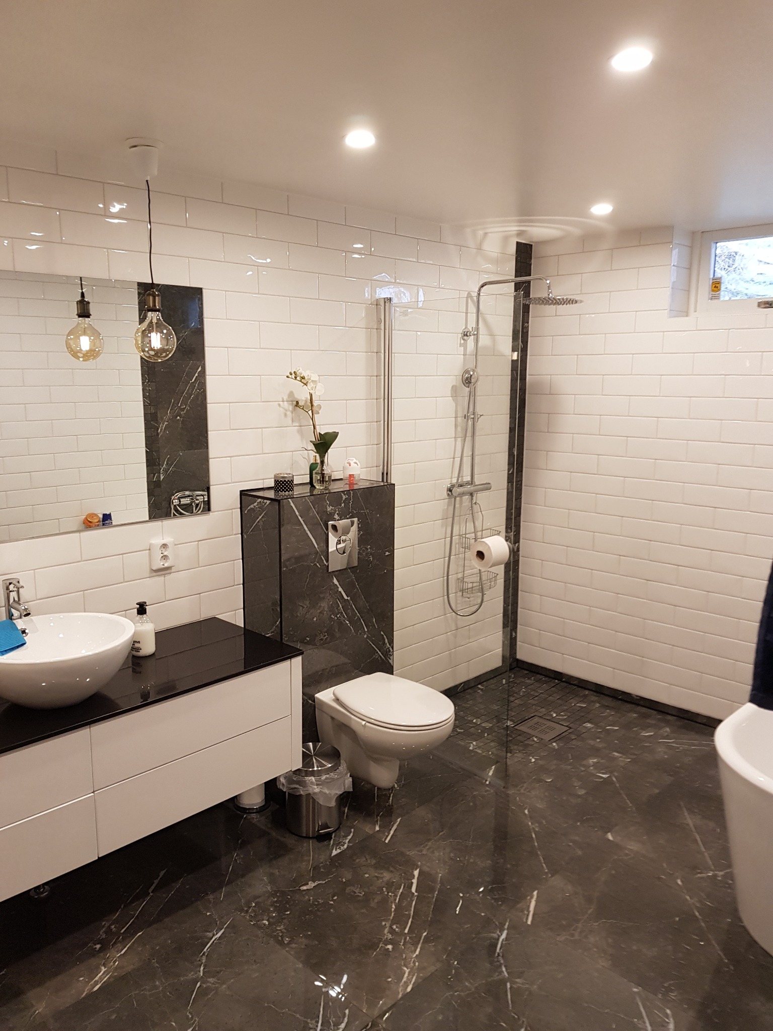 Nowoczesna łazienka z białymi kaflami, marmurową podłogą i akcentami w kolorze czarnym, z kabiną prysznicową typu walk-in i wiszącą toaletą.