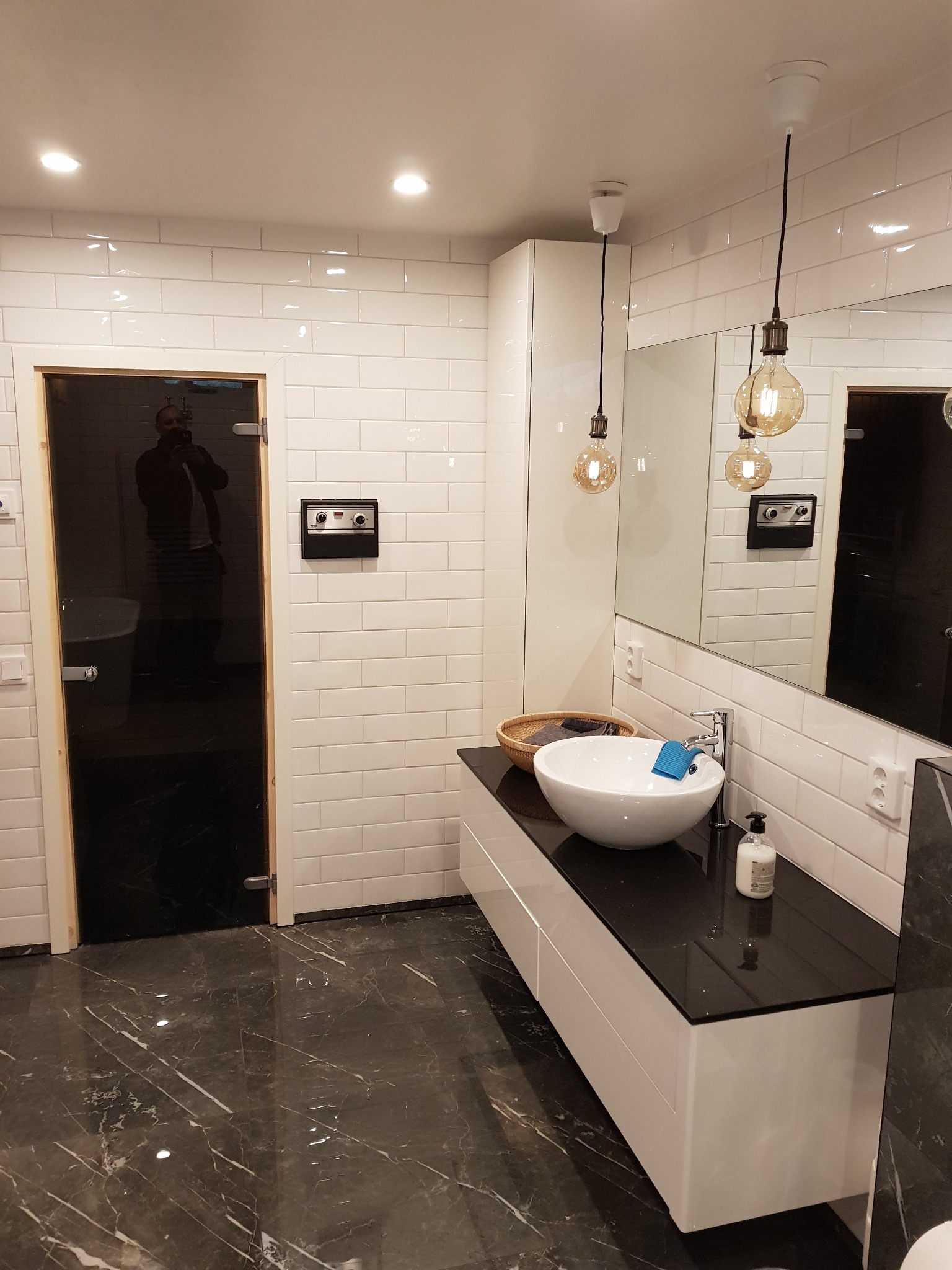 Nowoczesna łazienka z białą umywalką nablatową, czarnym blatem i marmurową podłogą, oświetlona wiszącymi żarówkami Edison, z widocznym odbiciem osoby robiącej zdjęcie w czarnych drzwiach.