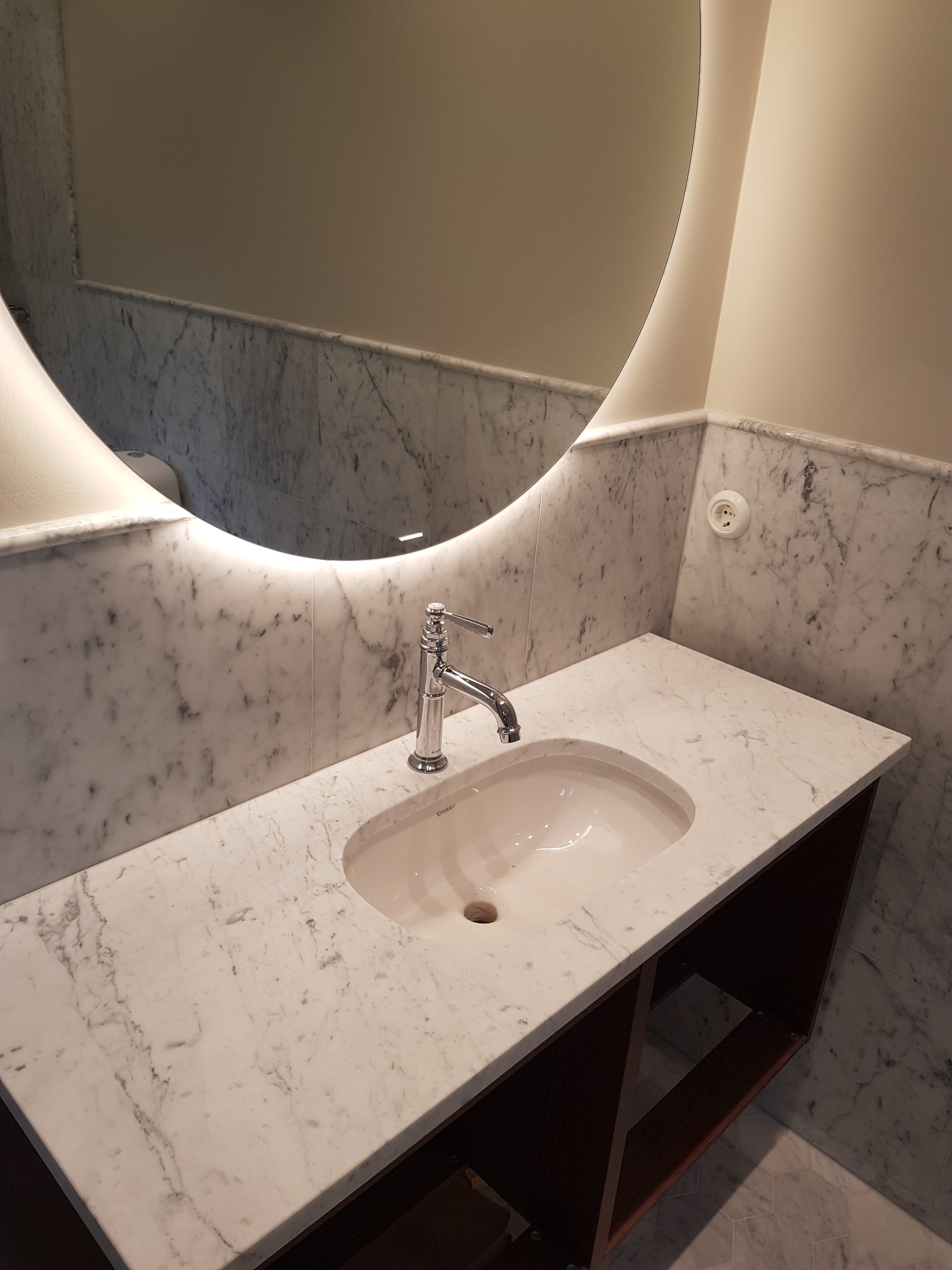 Umywalka Duravit wpuszczona w marmurowy blat z szafką, okrągłe lustro z podświetleniem LED, marmurowe ściany i gniazdko elektryczne.