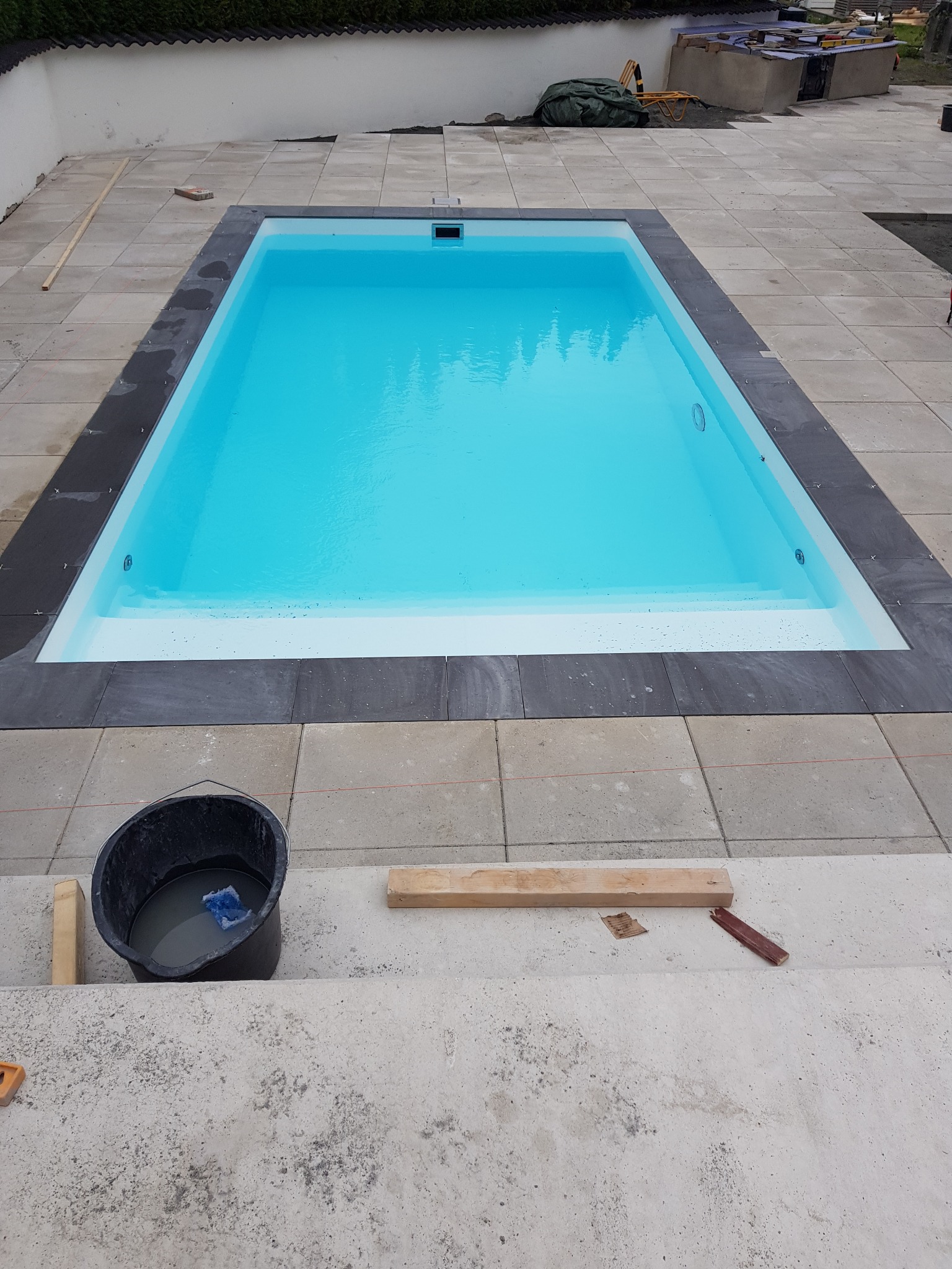 Nowy basen z błękitną wodą, obłożony jasnymi płytami chodnikowymi z ciemnoszarą obwódką, widok na etap prac wykończeniowych z wiadrem i narzędziami na pierwszym planie.